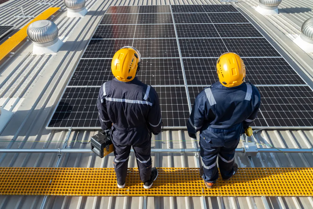 Vista aérea de ingenieros supervisando módulos solares negros en el tejado de metal de una fábrica o almacén.