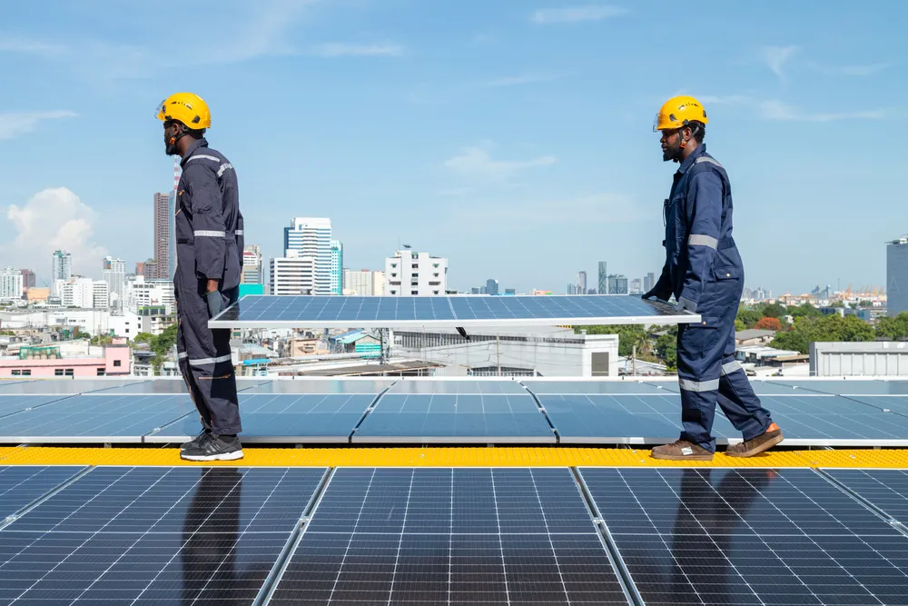 El trabajo en equipo es esencial en la instalación de paneles solares urbanos; dos trabajadores llevan un módulo bajo la luz del sol para generar energía limpia.
