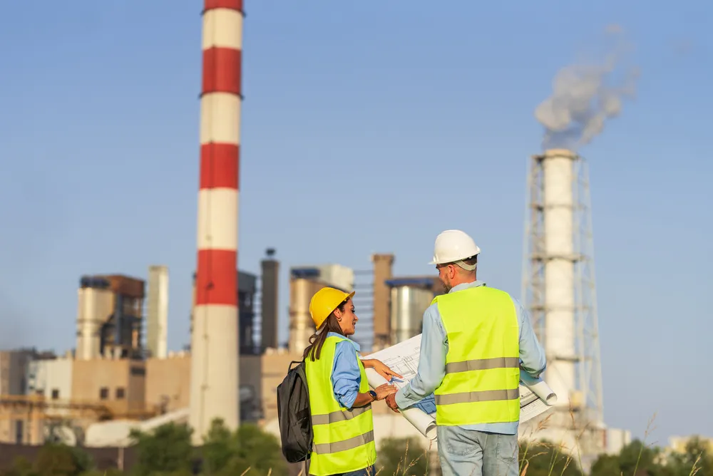 Dos ingenieros discuten los planos de la planta de energía con chimeneas humeantes al fondo, subrayando la gestión ambiental y el cumplimiento de los criterios ESG.