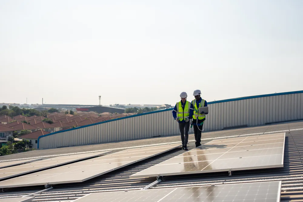 Dos ingenieros con cascos de seguridad y chalecos reflectantes revisan las instalaciones solares en una azotea industrial.