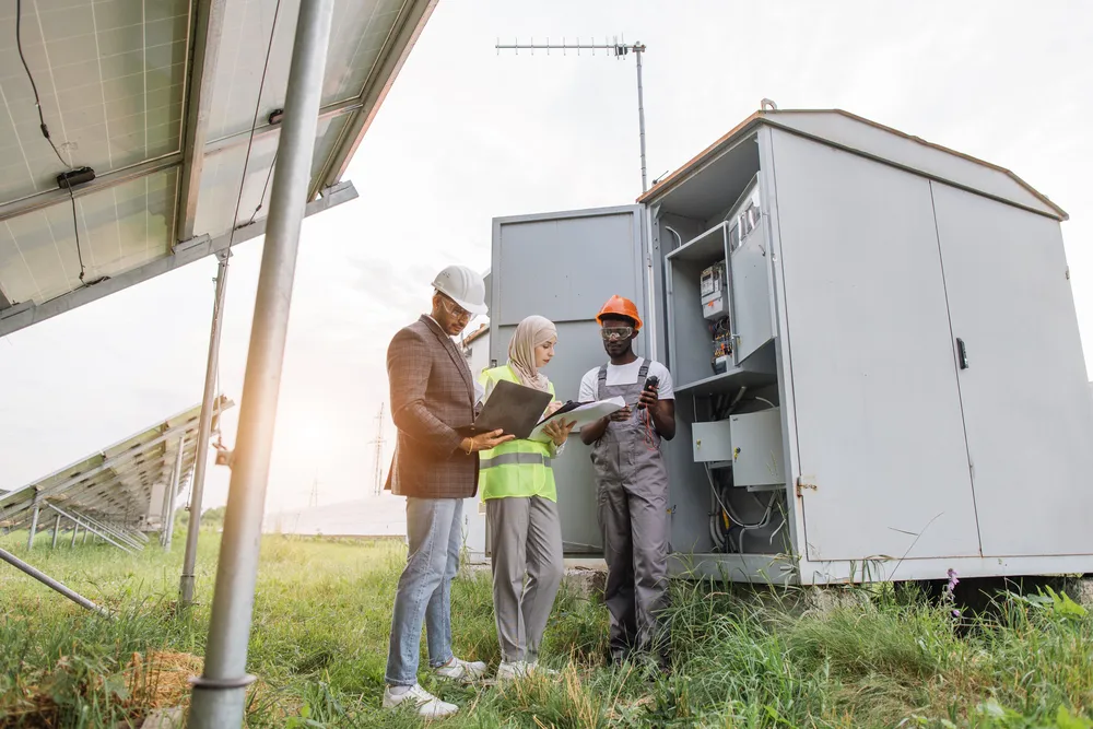 Equipo multirracial de técnicos inspecciona el gabinete de control de micro redes solares junto a paneles fotovoltaicos.