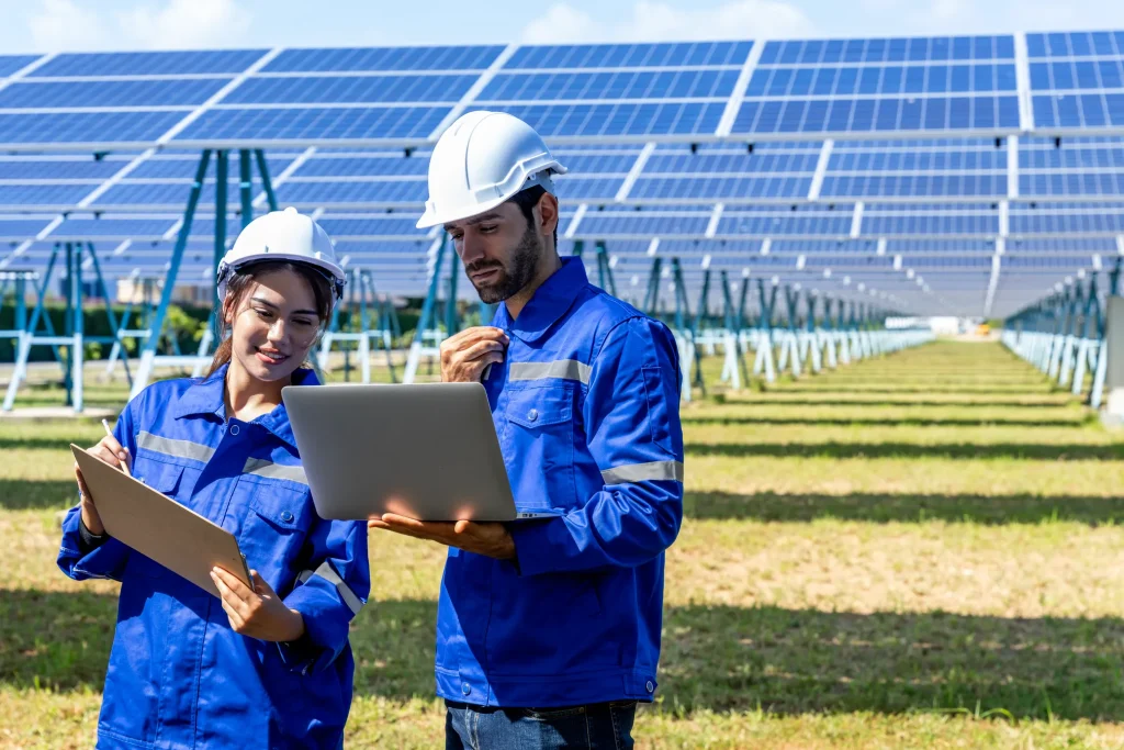 Qué es la certificación NABCEP: Una mujer y un hombre con uniformes azules operando una computadora portátil frente a filas de paneles fotovoltaicos