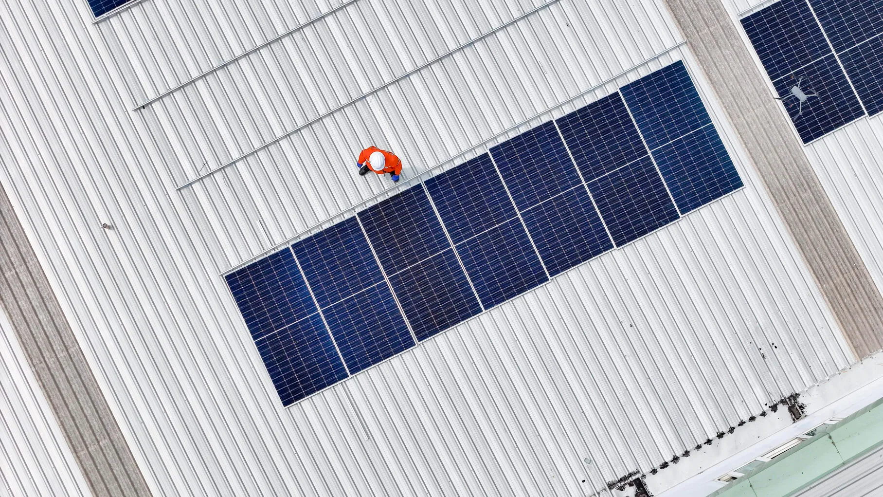 Operario en tejado metálico revisando la hilera de paneles solares para fábricas y naves industriales con un dron.