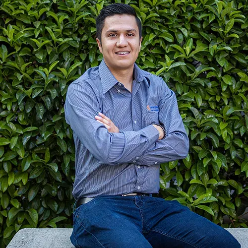 Luis Valadez, encargado de trámites en una empresa de paneles solares, sentado frente a un seto de hojas verdes.