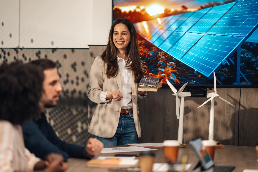 Una mujer sonriente presentando modelos de qué es la energía limpia y renovable ante una gran pantalla en una oficina.