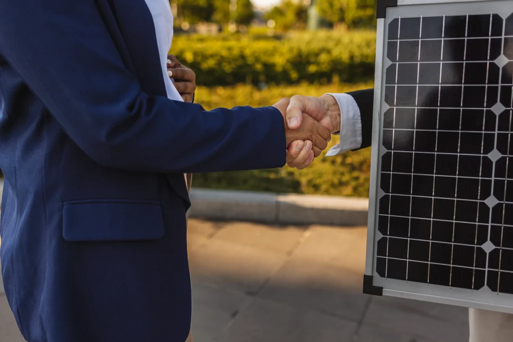 Primer plano de dos manos estrechándose en un acuerdo comercial, una de las personas sostiene un pequeño panel solar como símbolo de un trato sobre energía limpia.
